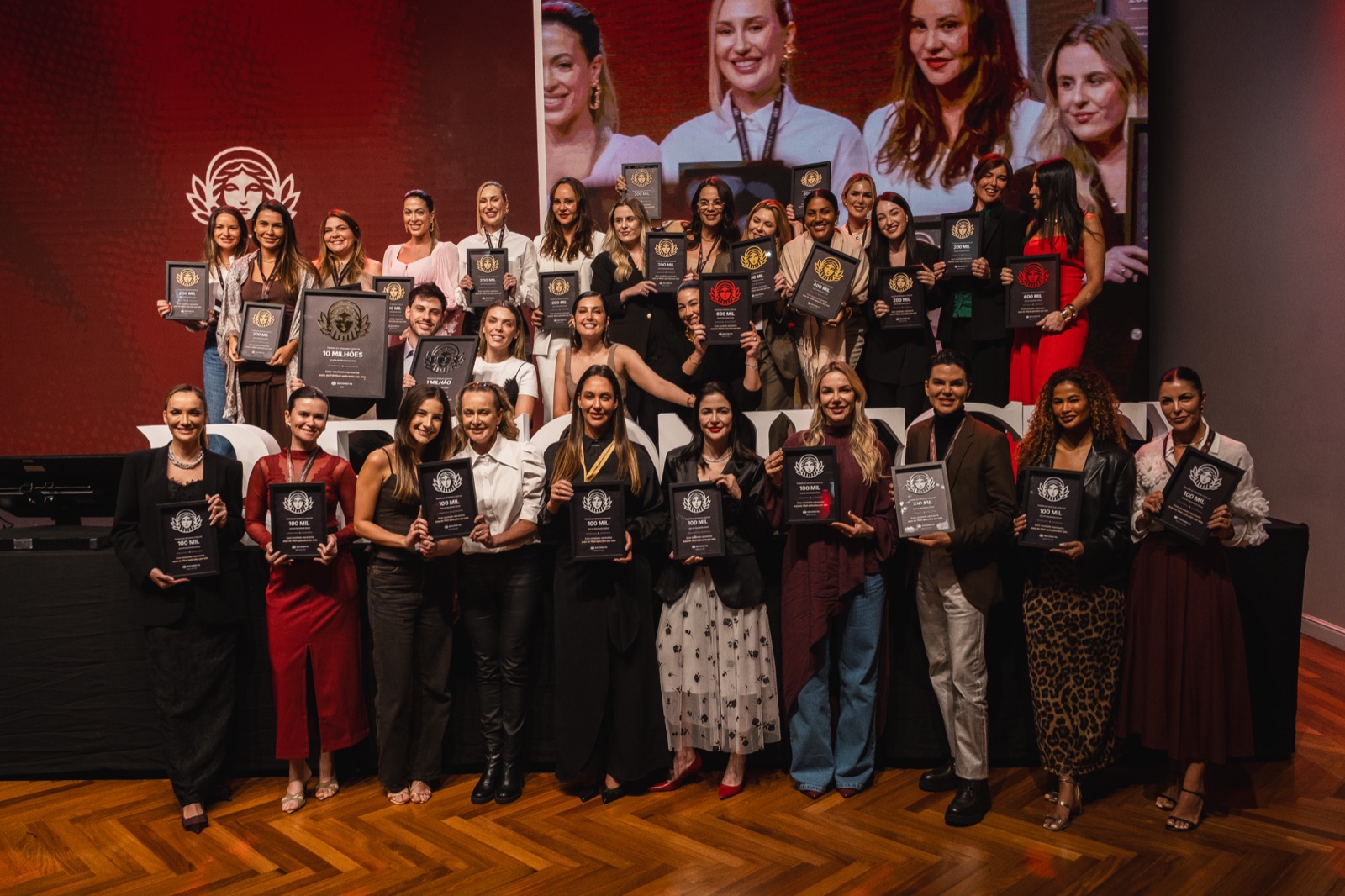 Grupo de mentoradas erguendo troféus no palco da premiação