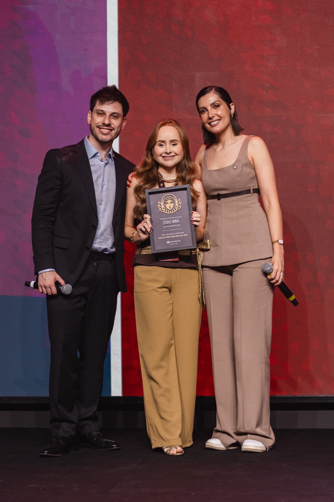 Mentorada recebendo troféu entre dois palestrantes no palco